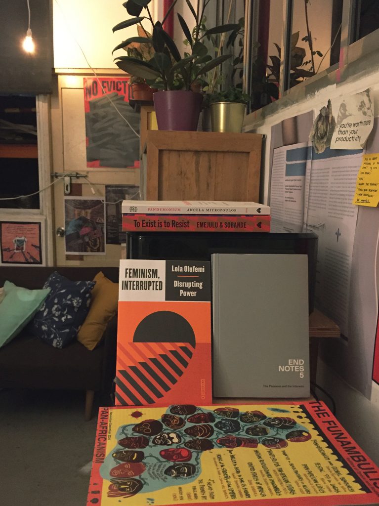 Photo of the library in warm lighting showing shelves, plants, a couch, and featuring some publications in the foreground, including "Feminism, Interrupted" by Lola Olufemi, End Notes 5, The Funambulist #32 on Pan-Africanism, "To Exist is to Resist" by Emejulu & Sobande, and "Pandemonium" by Angela Mitropoulos