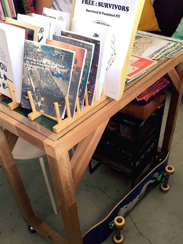 photograph of a display table in the library, zines and magazines can be seen on it, and a skateboard sits on the floor beside