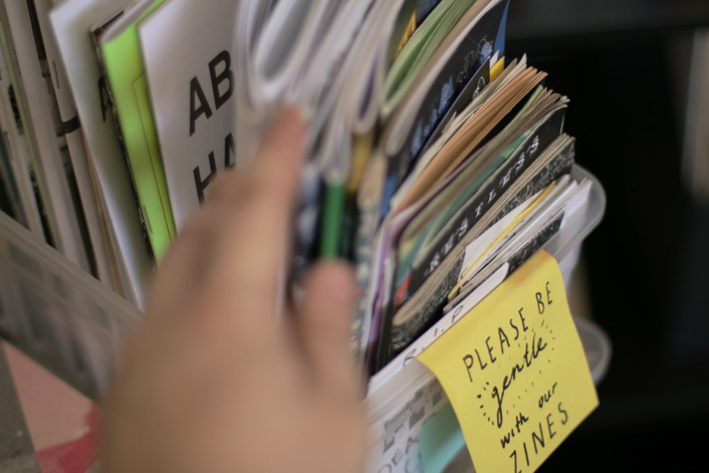 a close-up photo of a hand sifting through a plastic container with colourful zines standing in it. a handwritten post-it note on the front reads: "please be gentle with our zines"