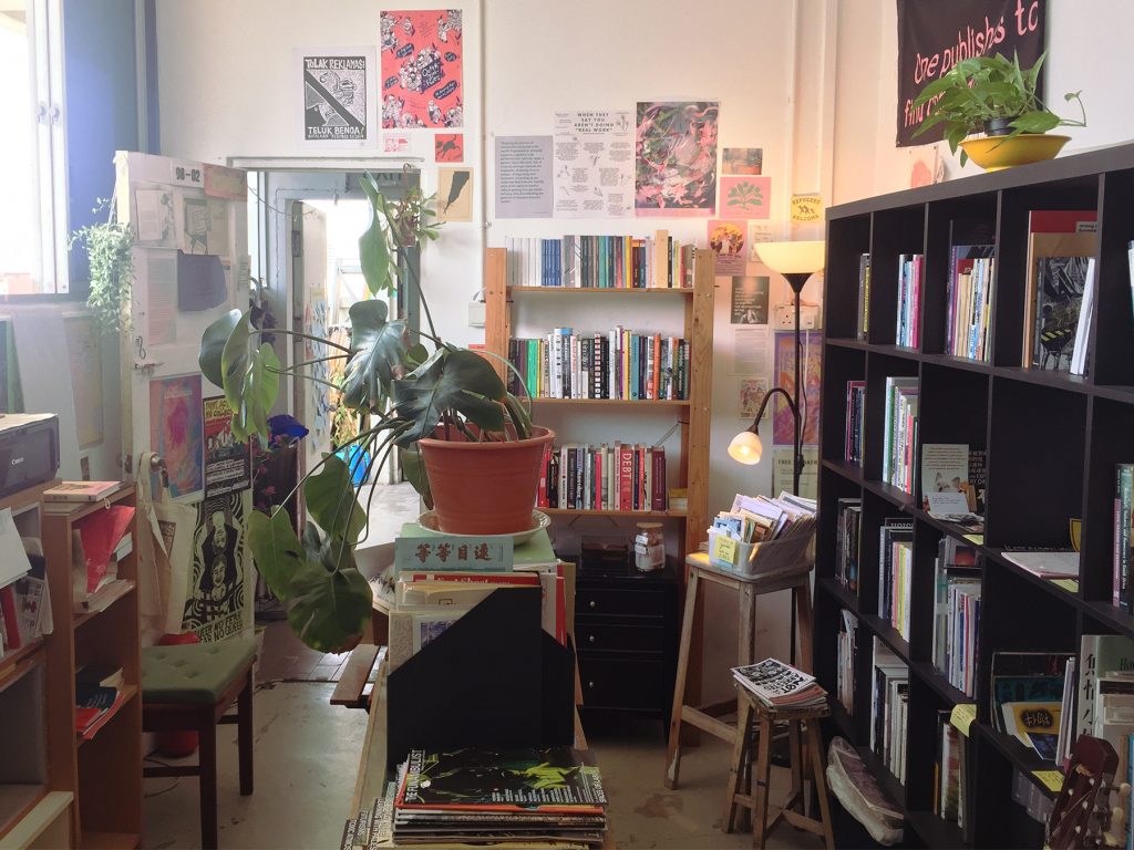 A photograph of the old wares infoshop library room in Geylang. Windows on the left and a doorway in front lights the room with daylight, there are posters on the walls and books on shelves, including a central island with magazines in stands. A large potted monstera plant also sits here, in the centre of the room and picture.