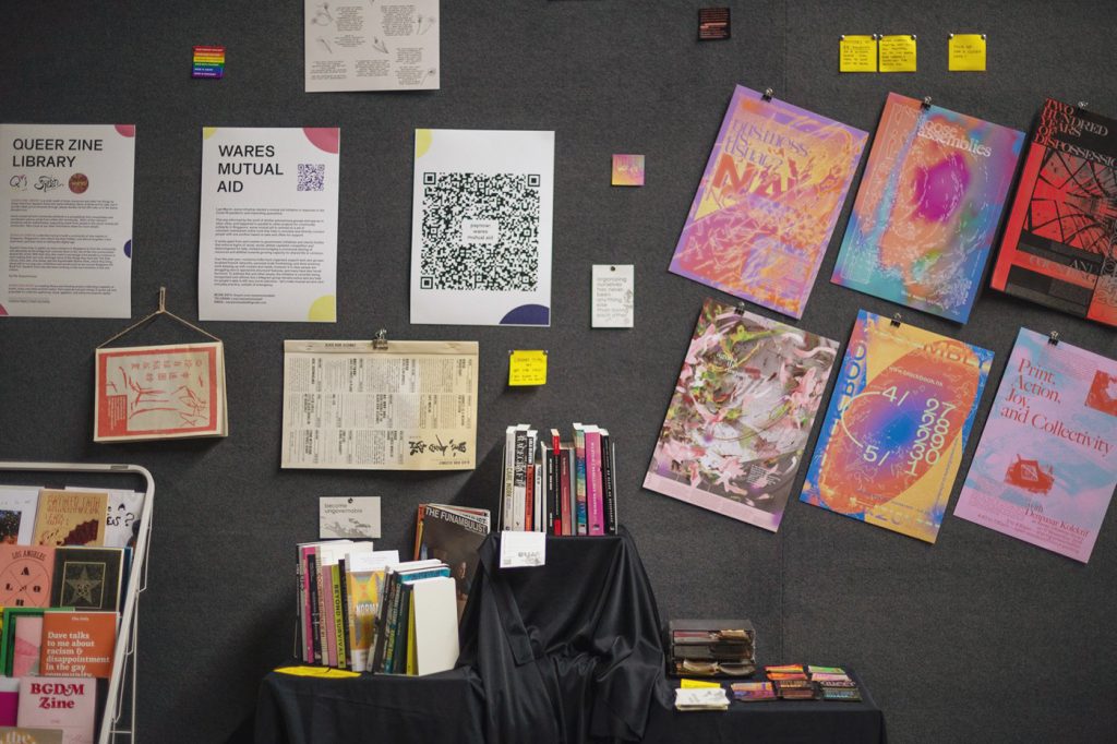 Photo of the wares set up at the Queer Zine Room in Singapore Art Book Fair 2021, which is directly against a dark grey carpet-lined wall. In the bottom middle are three tiers of makeshift display shelving covered in black velvet cloth, sitting on that is an assortment of books and magazines from the library standing upright and stickers on display. On the wall above and to the left are a series of notice texts for the Queer Zine Library and wares mutual aid, which proceeds from donations were going to. To the right of that are our six colourful posters hung diagonally in a grid. Scattered between all these are various sticky notes, flyers, and other publications for reading.