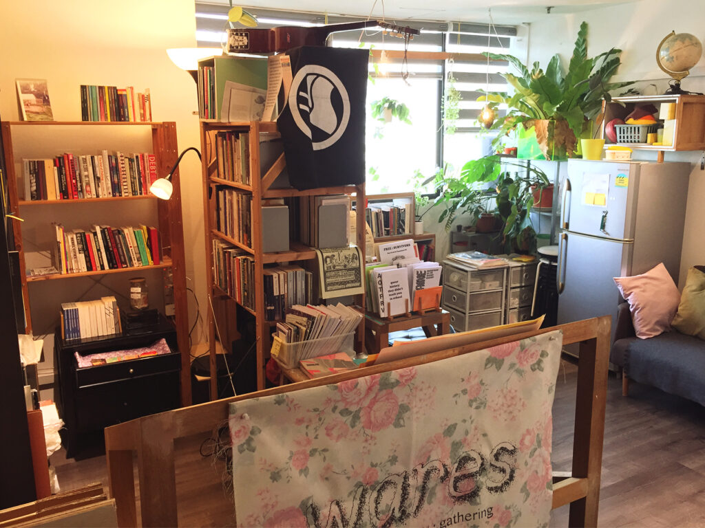 A photograph of the wares infoshop library room. It is framed at a slight angle, facing a wall with windows. The room is lit in warm yellow lighting. Wooden shelves with multiple tiers of books occupy the left half of the image, with boxes of zines visible as well, alongside other zines on display. At the top of a shelf in the middle of the frame is a black and white antifascist flag hanging from the top, with a classical guitar resting flat above it. On the right half of the image, flanking the windows are more shelves and potted plants, including a large leafy bird's nest fern and monstera. There is a fridge and and sofa as well. In the foreground, a floral patterned fabric banner pinned on a display shelf reads "wares".