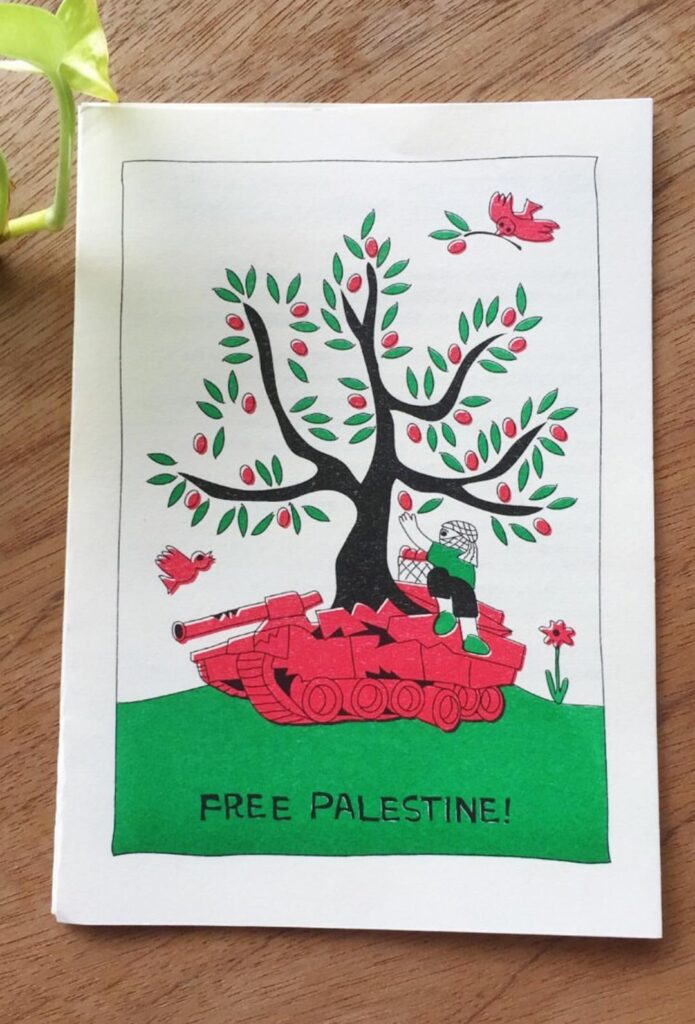 Photo of a stylised and simple drawing printed with 3 colours: red, green, and black, on off-white paper. It features an olive tree growing out of a wrecked tank sat on a mound, its tracks and barrel broken. A childlike figure, head wrapped in a keffiyeh, sits on the tank, they reach out picking the olive fruits, placed into a basket beside them. A flower blooms nearby, and two birds fly free around the tree, one with an olive branch and fruit in beak. At the bottom of the drawing, handwritten with all uppercase letters: “FREE PALESTINE!”